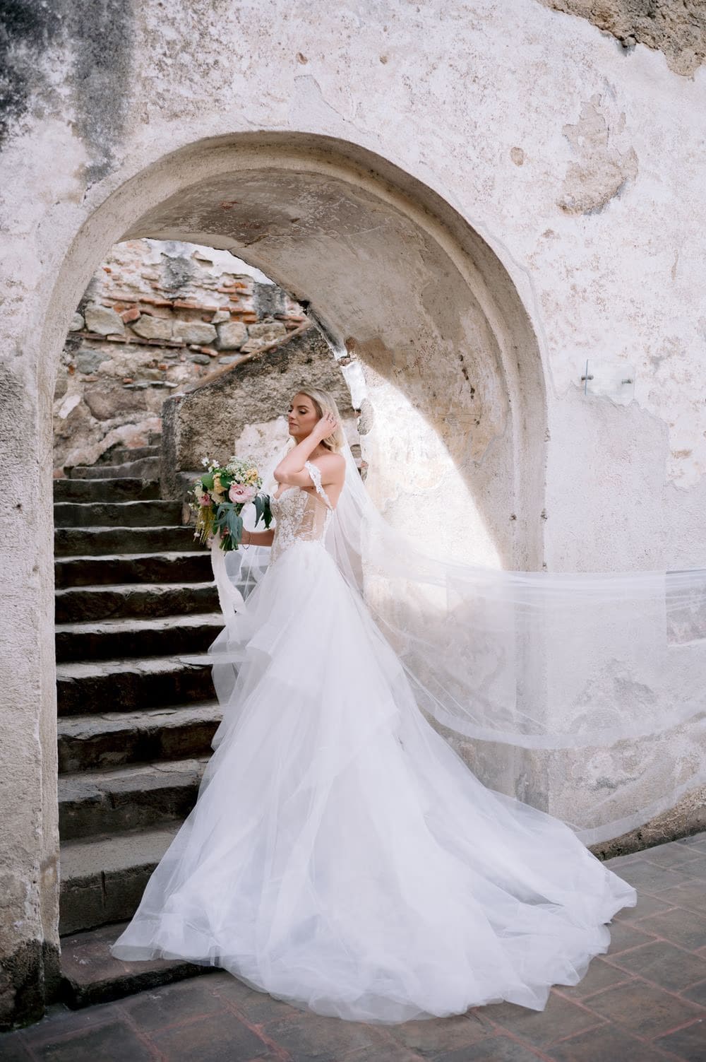 Bride stands quietly beneath a stone arch at La Recolección Ruins, bouquet at her side.