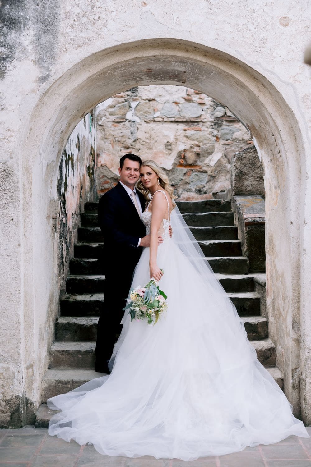 Sydney and Nathan pose under a stone arch with the gown's tulle train spread across the steps of La Recolección.