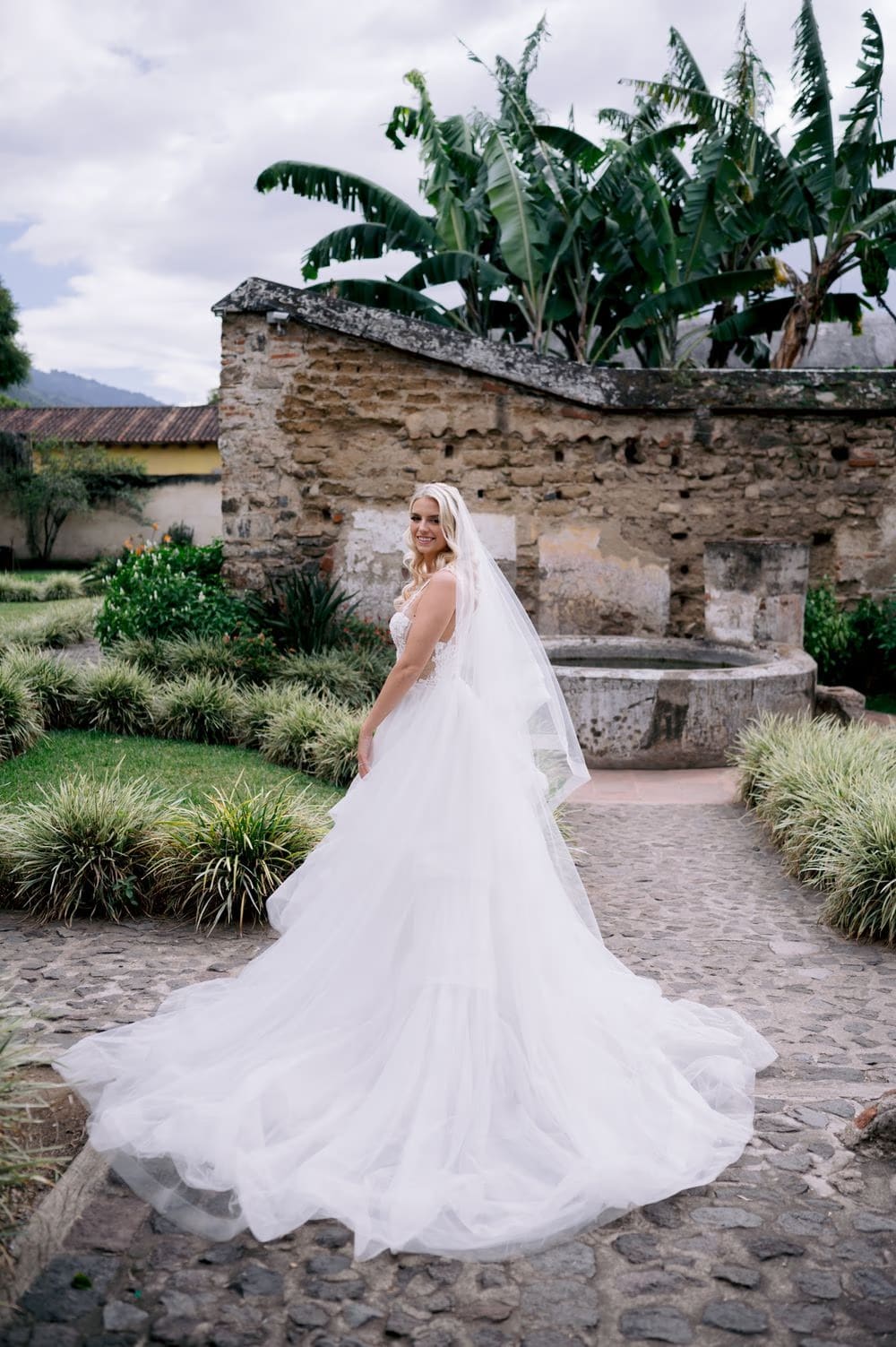 Bride turns in a garden courtyard with banana palms and an old stone fountain behind her in Antigua Guatemala.
