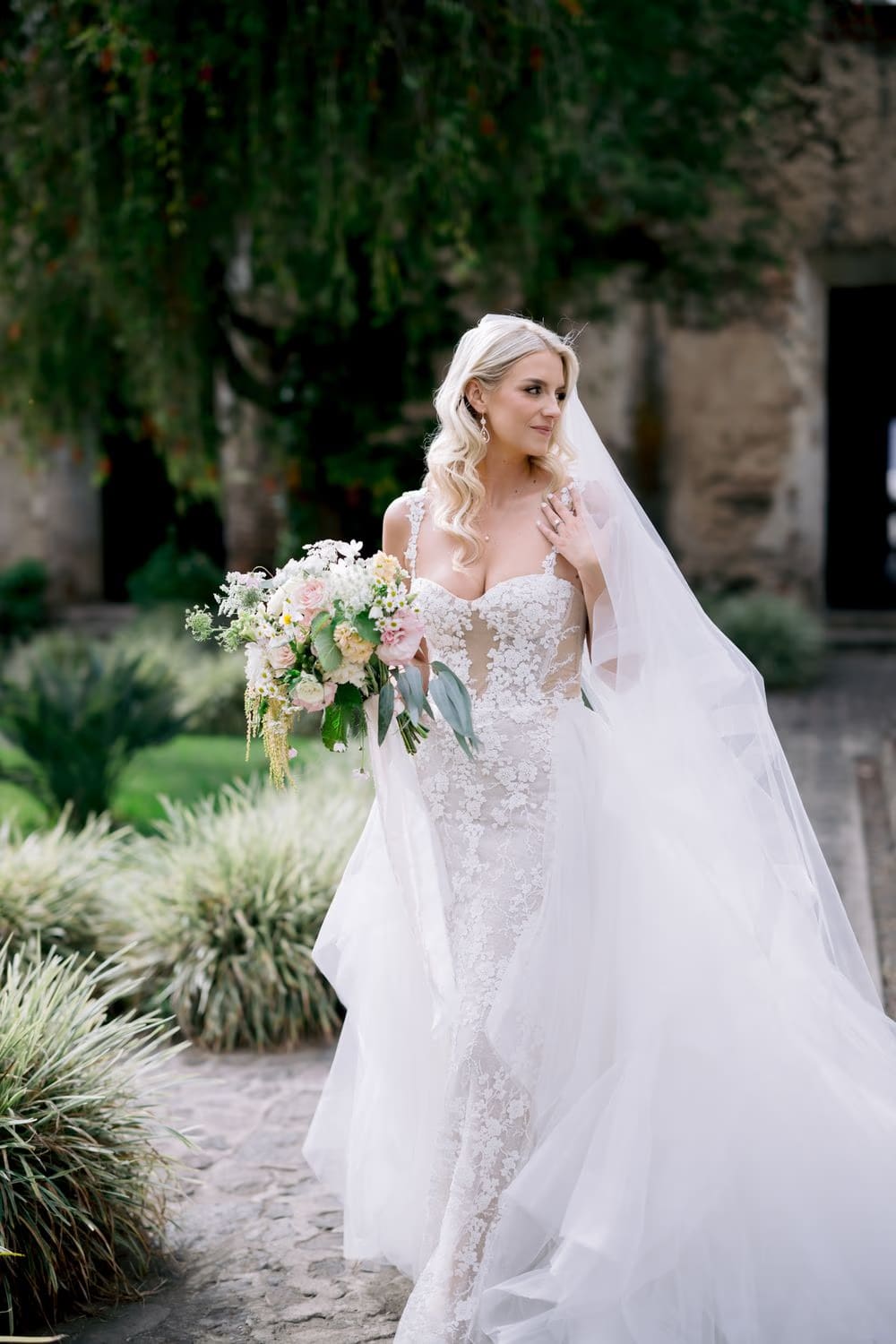 Bride Sydney holds her wild garden bouquet in a leafy courtyard with a stone wall behind — destination bridal portrait.