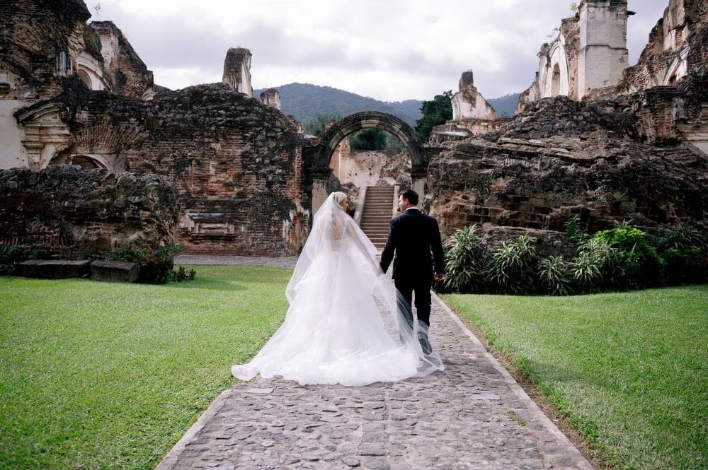 Bride and groom walk toward the iconic stone arch of La Recolección Ruins in black and white — destination wedding portrait in Antigua Guatemala.