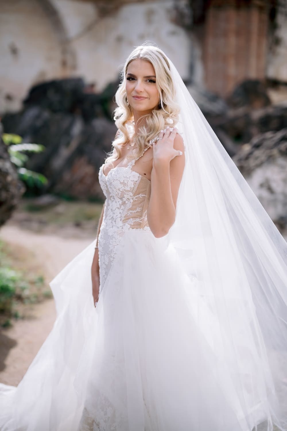 Bride Sydney touches her shoulder strap in a soft-lit ruin garden — editorial bridal portrait.