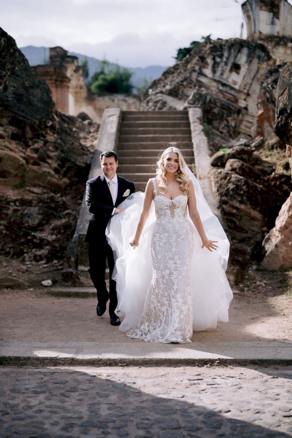 Bride leads her groom down the cobblestone steps of an Antigua street, smiling toward the camera — colonial Antigua wedding portrait.