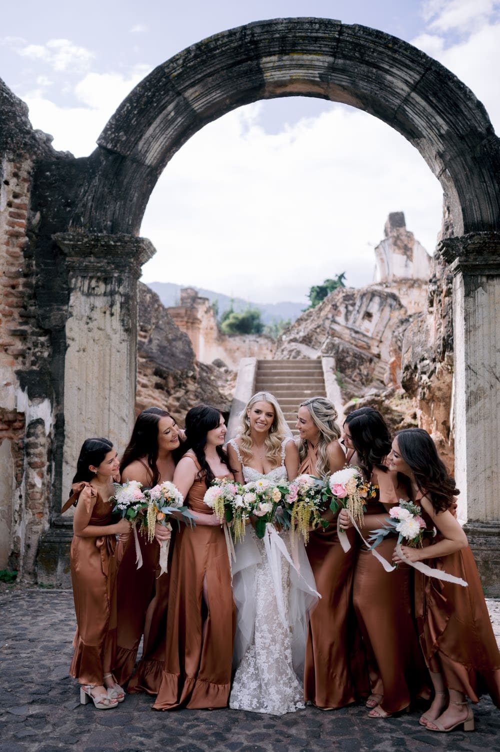 Bride and bridesmaids in copper satin gowns laugh together framed by the great arch of La Recolección Ruins.