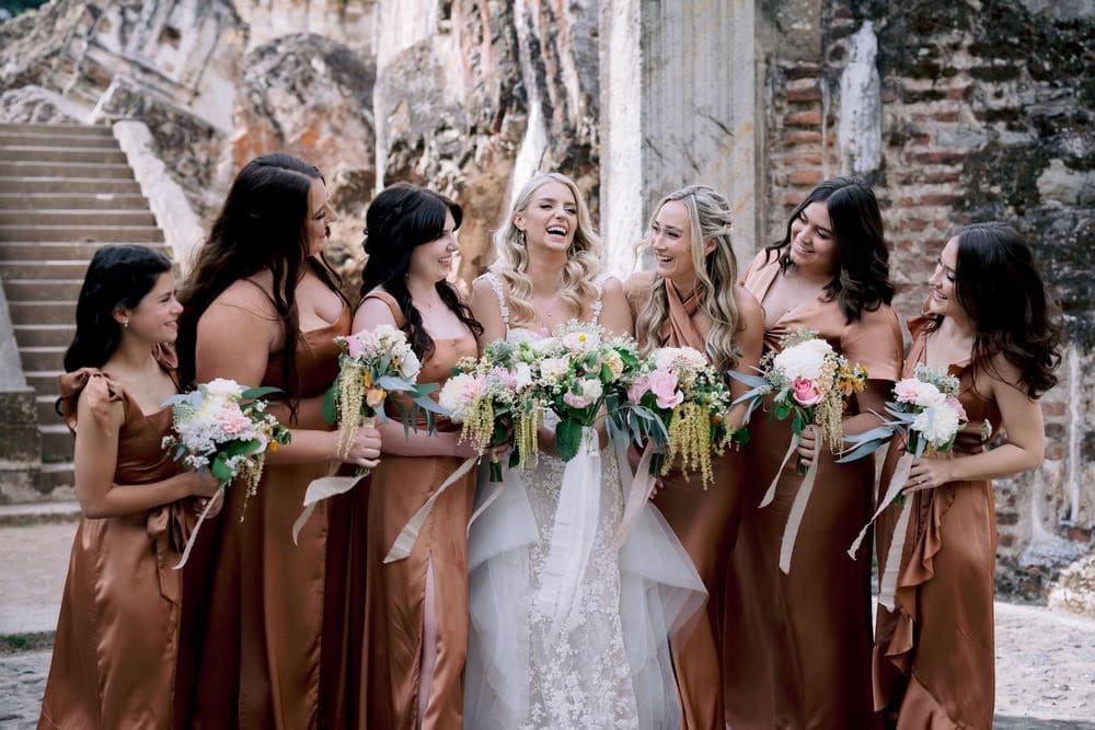 Sydney shares a laugh with her bridesmaids in copper satin gowns near the ruins of La Recolección in Antigua Guatemala.