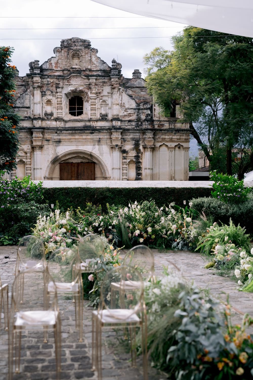 Garden ceremony aisle lined with wild florals and ghost chairs facing the San José el Viejo facade in colonial Antigua.