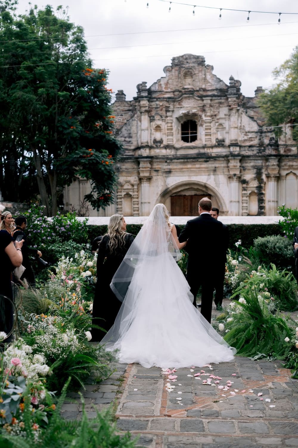 Bride walks down the petal-strewn aisle on her father's arm toward Nathan with the San José el Viejo facade ahead.