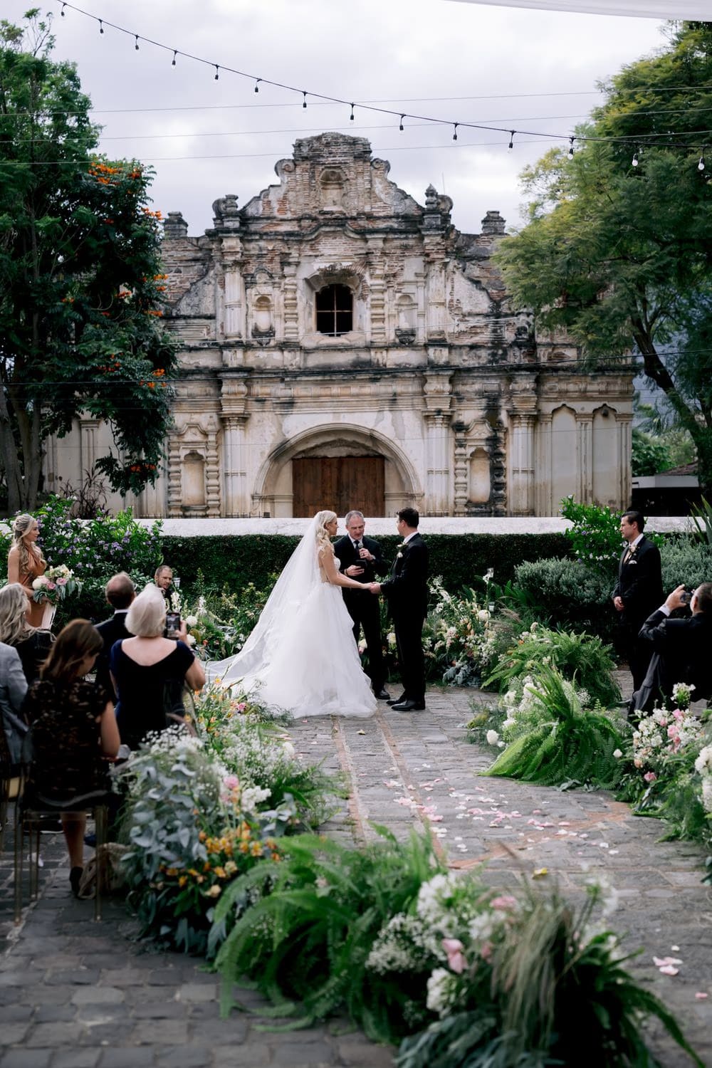 Sydney and Nathan hold hands during the ceremony with the floral aisle and San José el Viejo facade behind them in Antigua Guatemala.