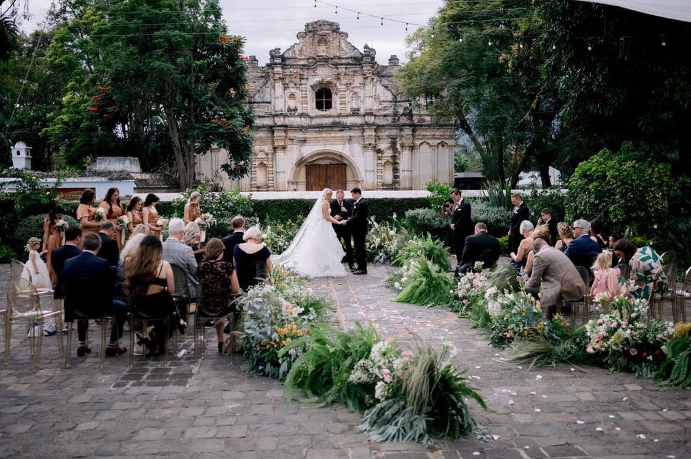 Wide-angle of the outdoor ceremony with seated guests, lush florals and the San José el Viejo facade as backdrop.