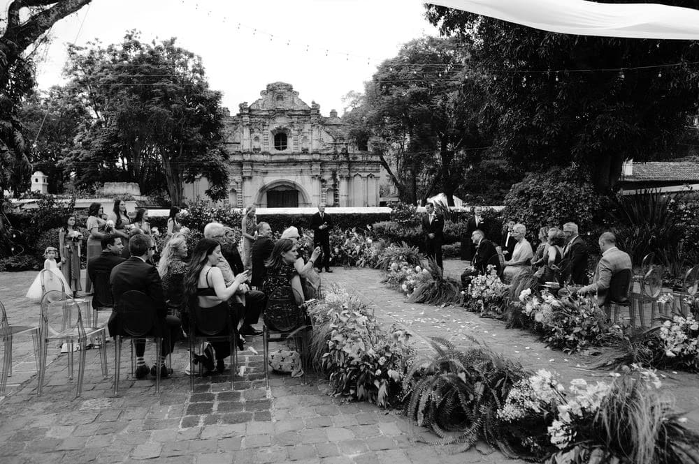 Black-and-white wide-angle of the ceremony at San José el Viejo with the facade rising behind the floral aisle.