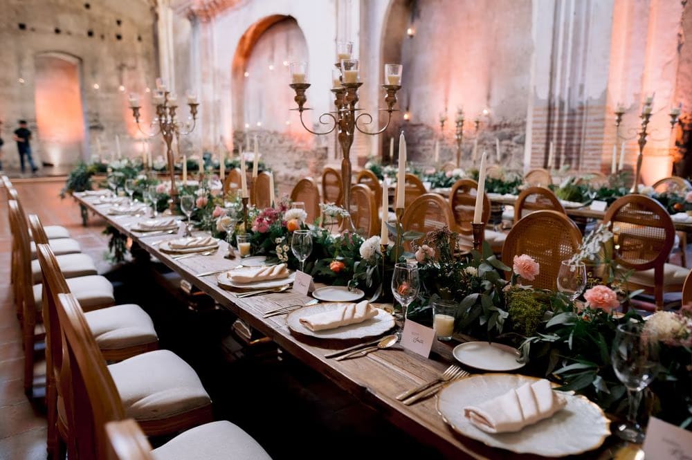 Long candlelit reception tablescape inside the nave of San José el Viejo, glowing warm against weathered ruin walls.