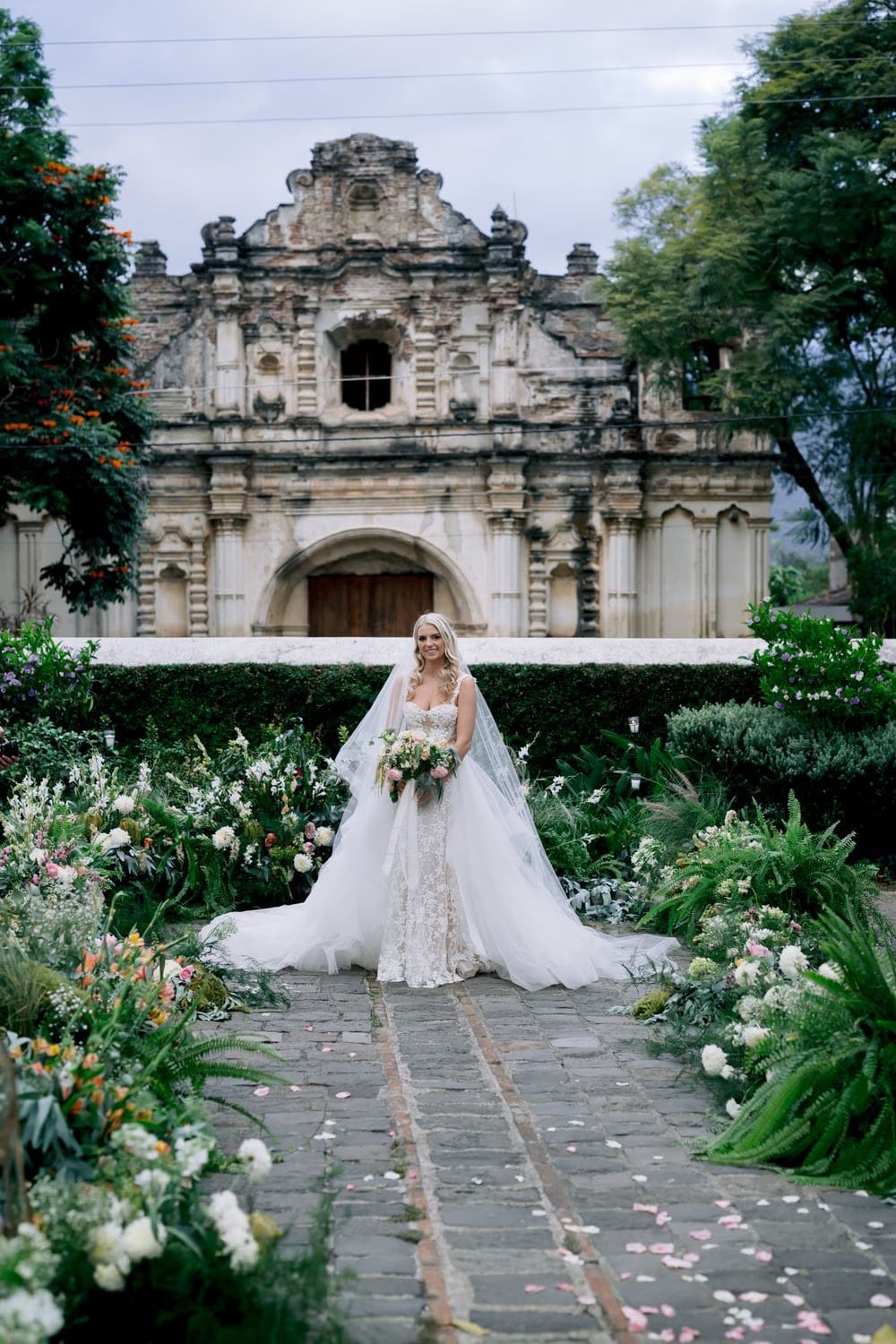 Bride Sydney stands on the petal-strewn aisle in front of the San José el Viejo facade in Antigua Guatemala — editorial bridal portrait.