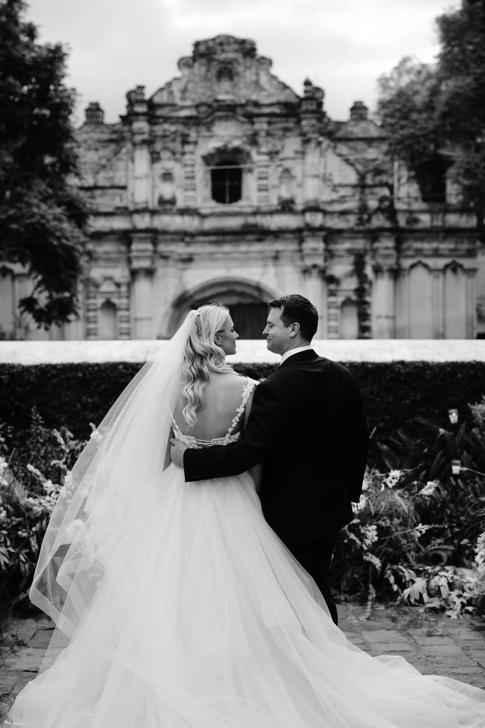 Bride and groom from behind in black and white facing the San José el Viejo facade — editorial wedding portrait.