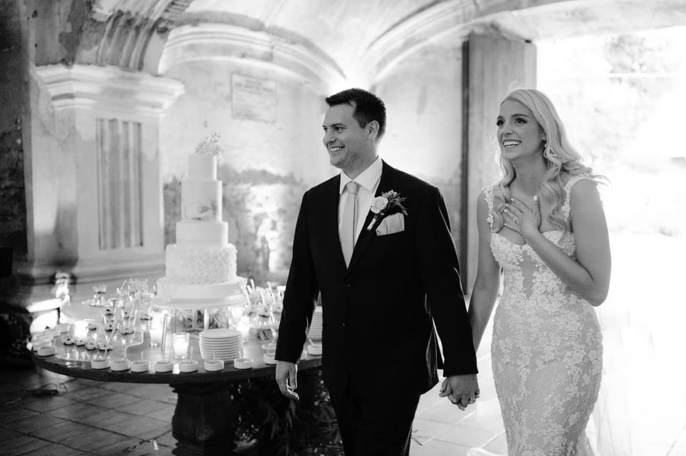 Sydney and Nathan walk toward their towering wedding cake during the reception at San José el Viejo, in black and white.