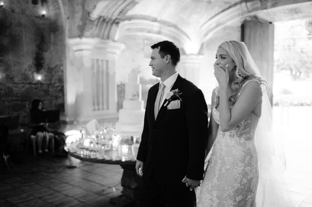 Bride covers her mouth in surprise as they take in their wedding cake at the San José el Viejo reception — black and white.