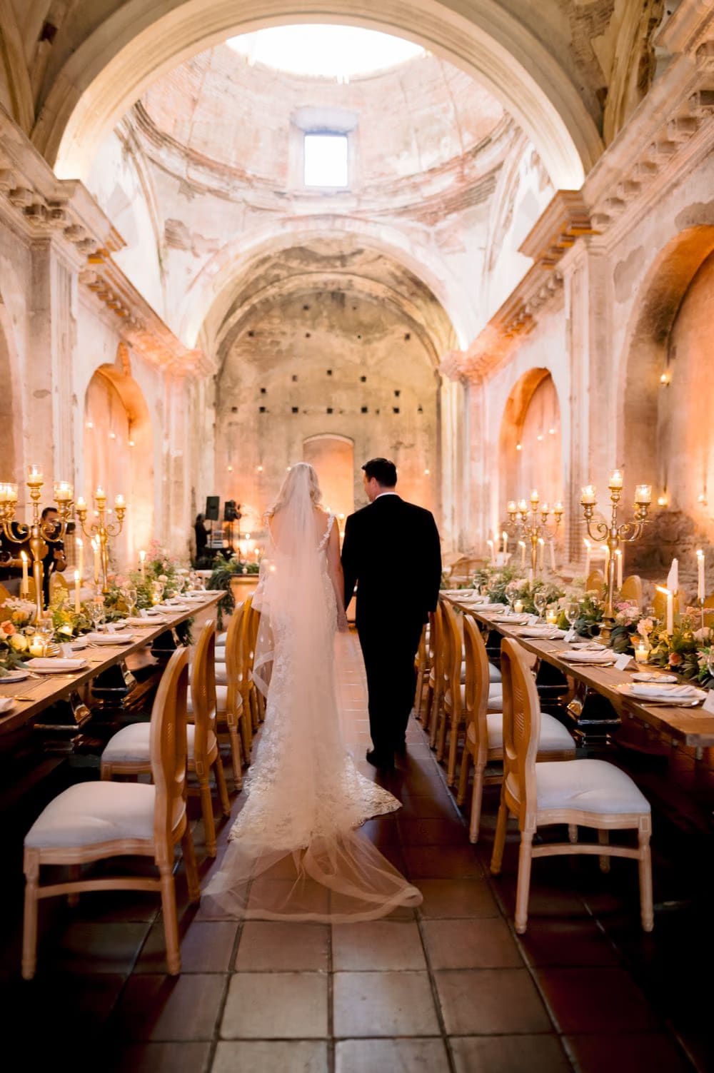 Bride and groom walk into the candle-lit nave of San José el Viejo Ruins between the long reception tables.