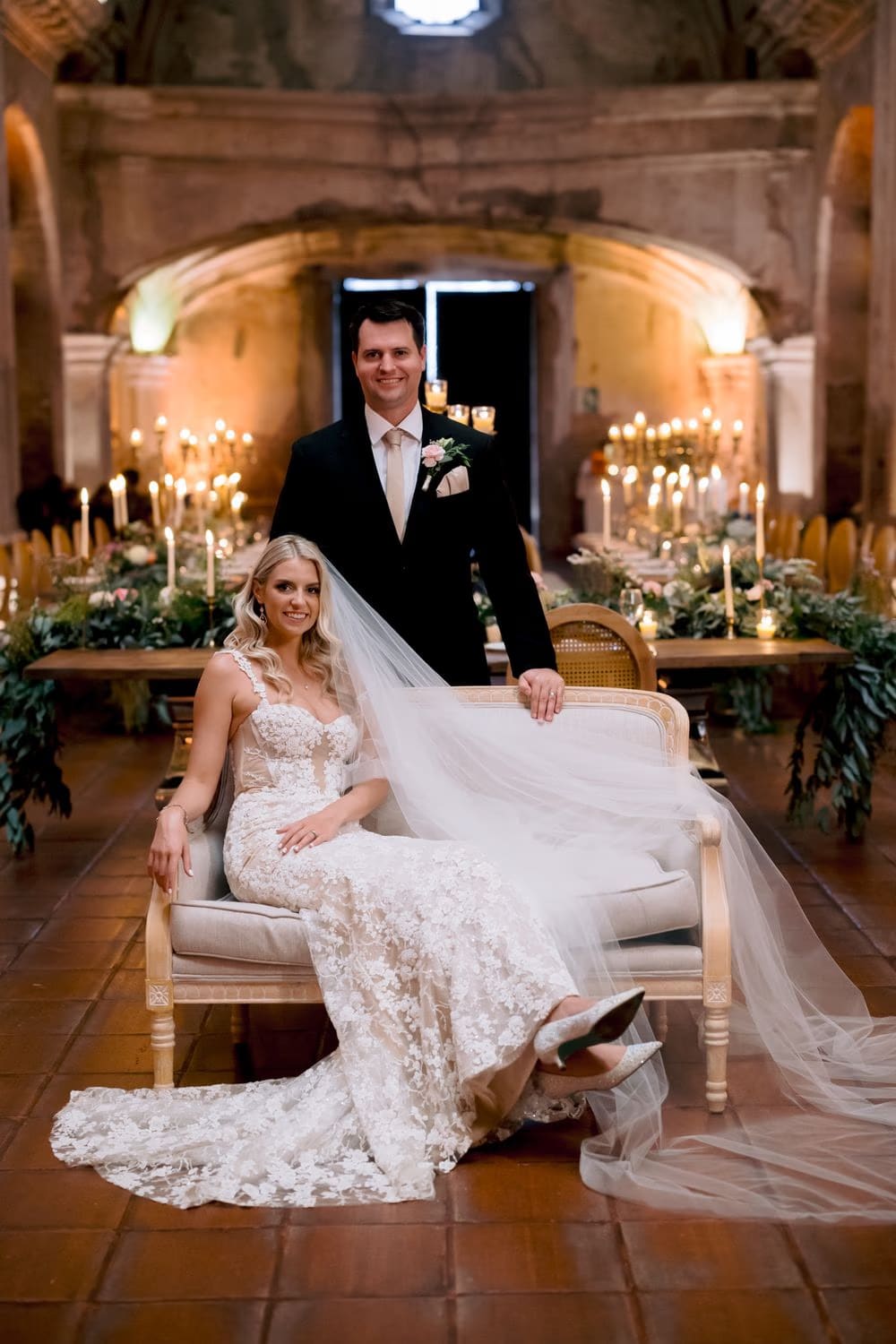 Sydney sits on a vintage sofa with Nathan standing behind her inside the candle-lit reception at San José el Viejo.