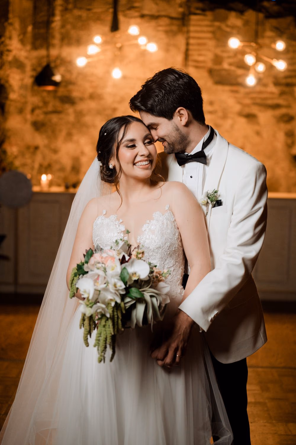 Bride holds her white orchid and amaranth bouquet by Addy Florales while the groom leans in against a string-light wall at Hotel Casa Santo Domingo.