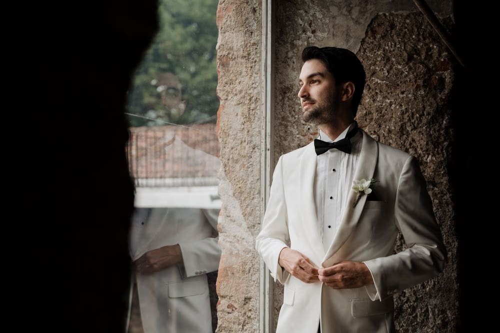 Groom buttons his white tuxedo jacket against a stone wall — elegant wedding portrait in Antigua Guatemala.