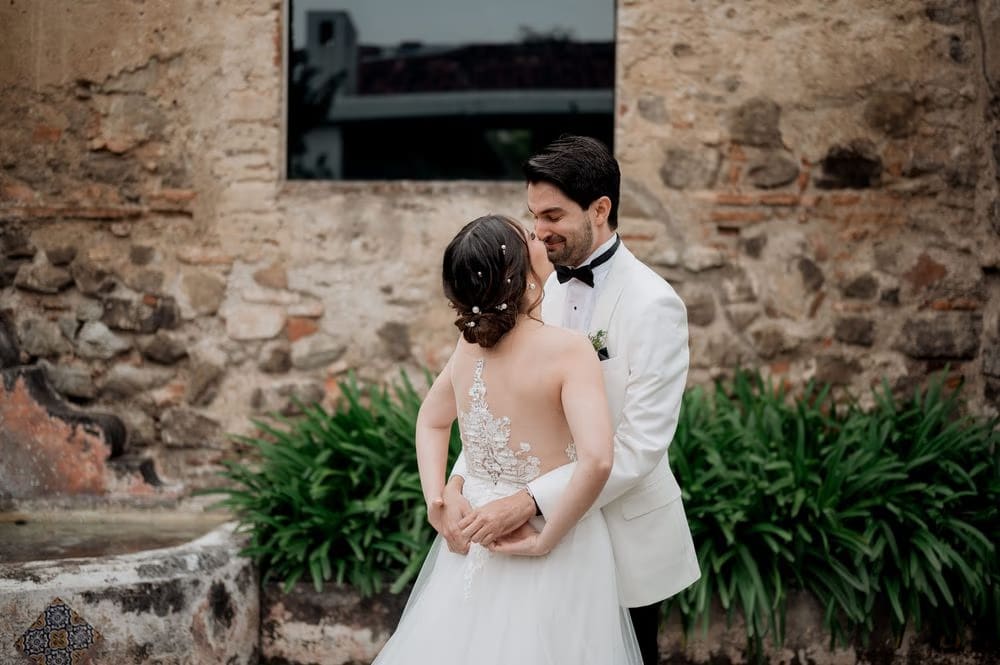 Bride and groom share a soft forehead kiss in front of an ivy-flecked stone wall in Antigua Guatemala.
