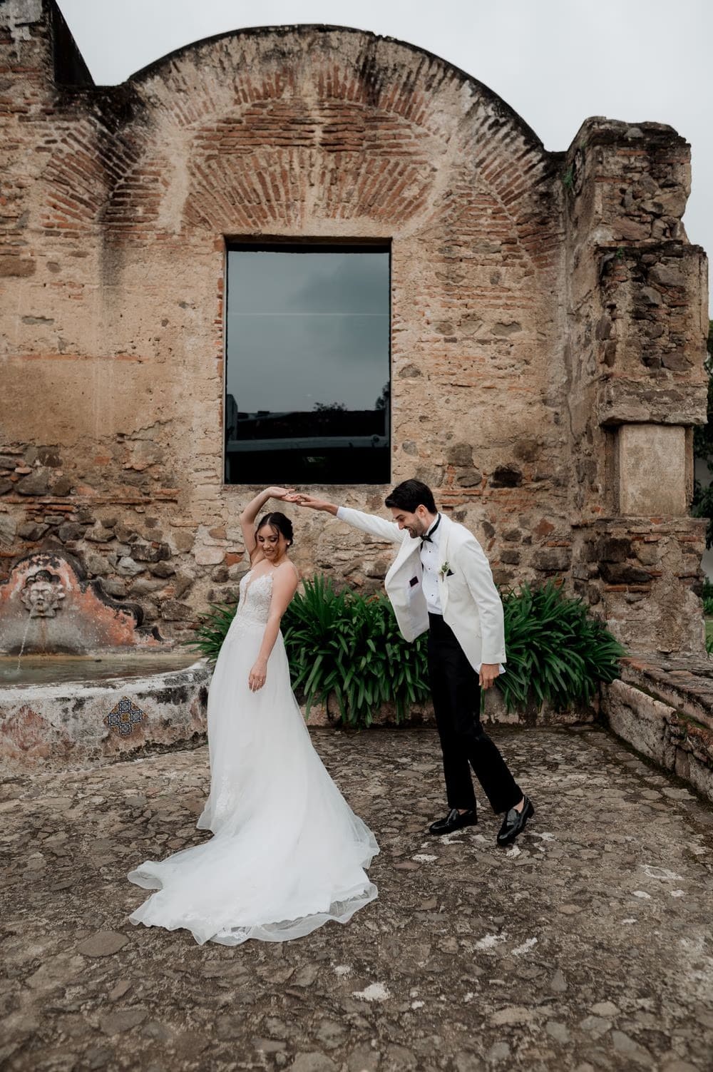 Groom twirls the bride beneath a brick arch ruin with stone fountain detail — destination wedding in Antigua Guatemala.