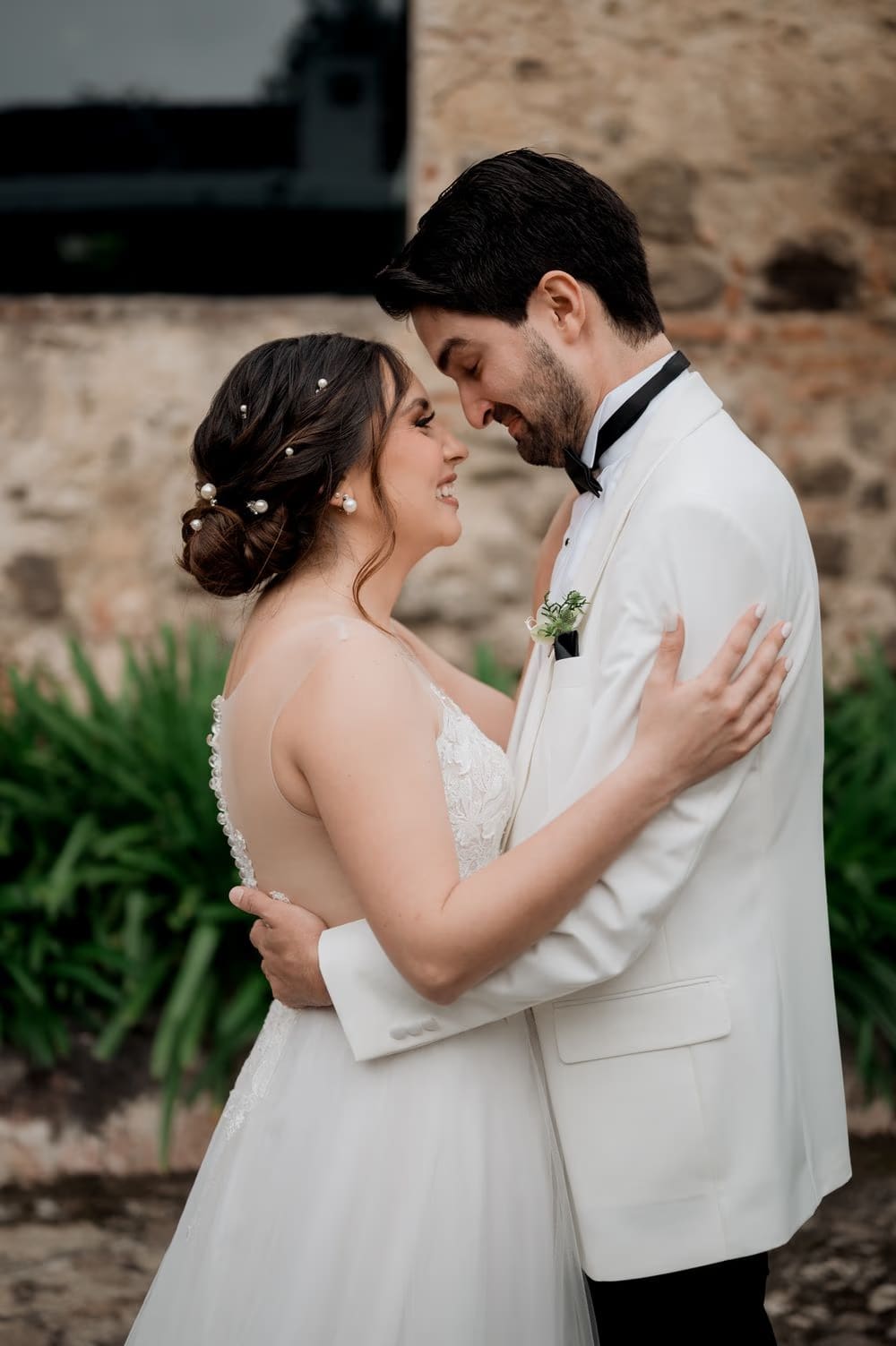 Bride and groom hold each other close at a stone wall, her pearled updo lit warmly — colonial monastery, Antigua.