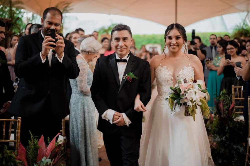 Bride walks down the aisle on her father's arm beneath the canopy as guests film and applaud — destination wedding in Antigua Guatemala.
