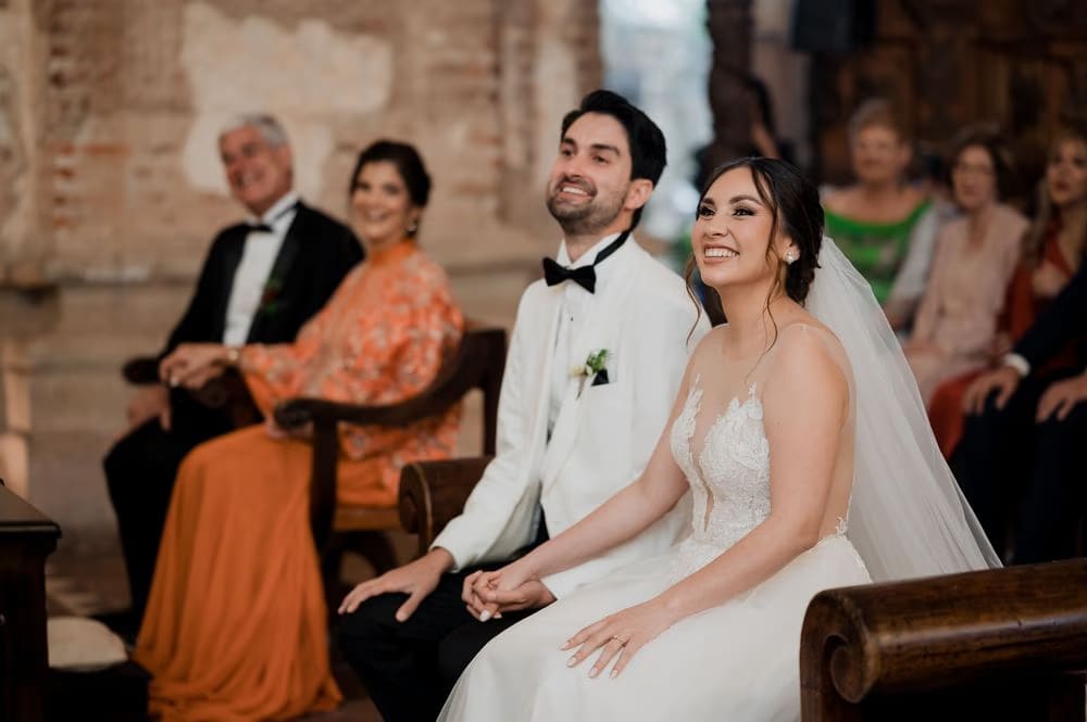 Bride and groom share a laugh in the pew during their wedding ceremony as parents look on at Casa Santo Domingo.
