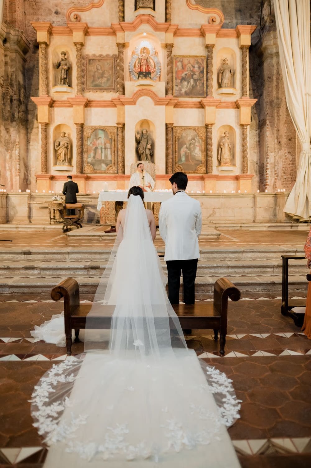 Wide back view of the couple at the baroque altar with the bride's lace cathedral veil trailing across the tile floor.