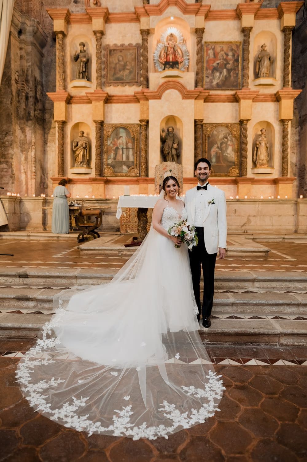 Bride and groom pose at the foot of the gilded altar with the lace-edged cathedral veil sweeping behind them.