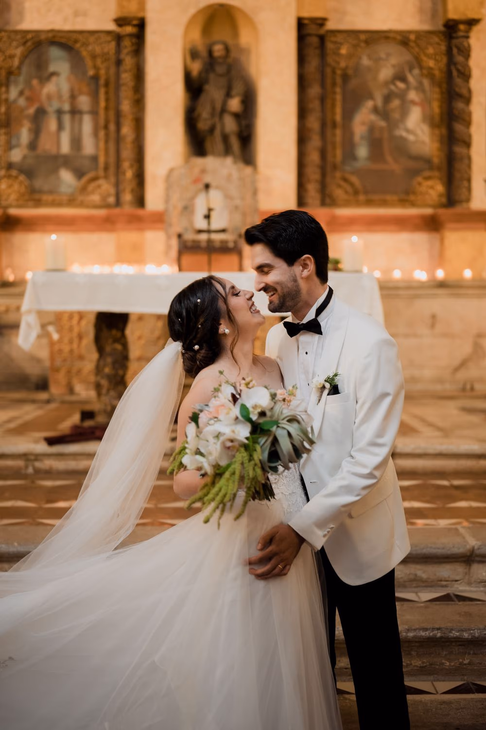 Bride and groom smile cheek-to-cheek before the candle-lit baroque retablo holding her cascading bouquet by Addy Florales.