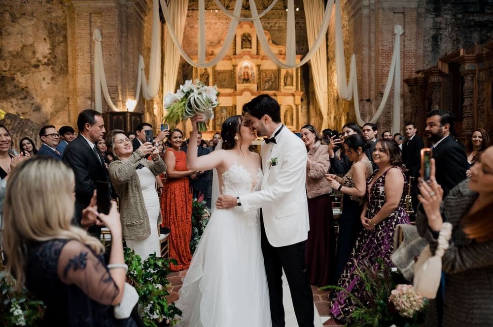Couple kiss mid-aisle under draped fabric and chandeliers as guests photograph the moment in Antigua Guatemala.