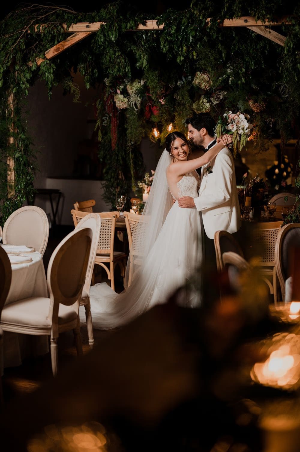 Bride beams over the groom's shoulder under a wooden pergola dressed in greenery and florals by Addy Florales.