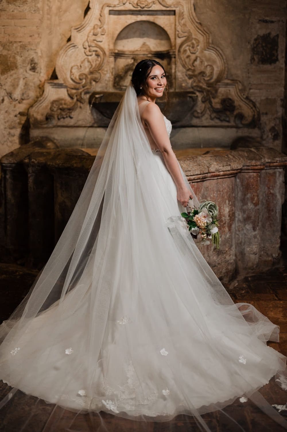 Solo bridal portrait with cathedral veil cascading on stone floor in front of a baroque carved niche.
