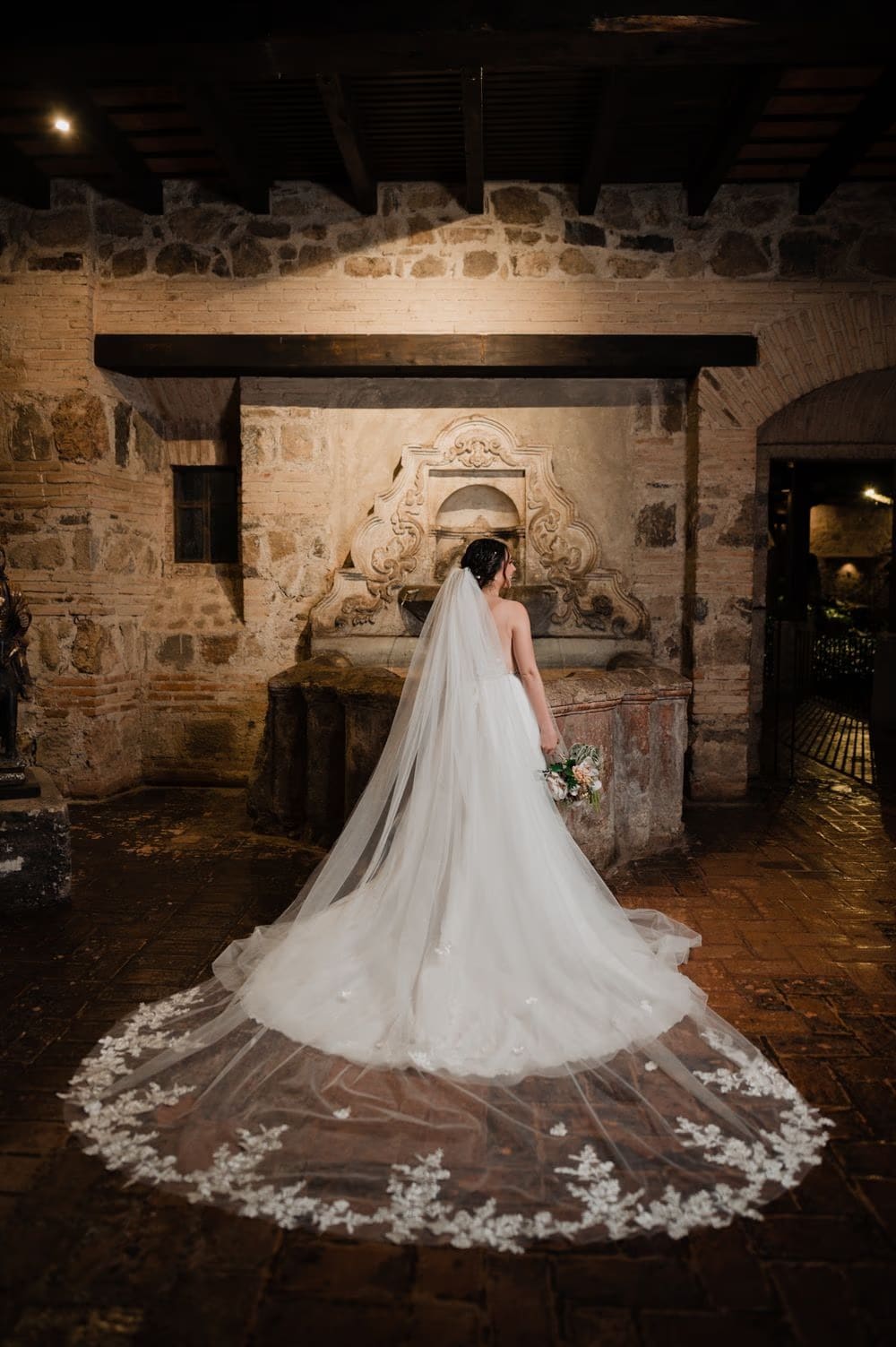 Back portrait of the bride with cathedral veil unfurled across brick floor by an old stone fountain at the monastery.