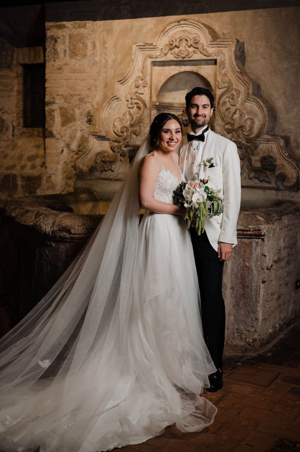Full-length newlywed portrait by the ornate stone niche with cathedral veil sweeping behind the bride.