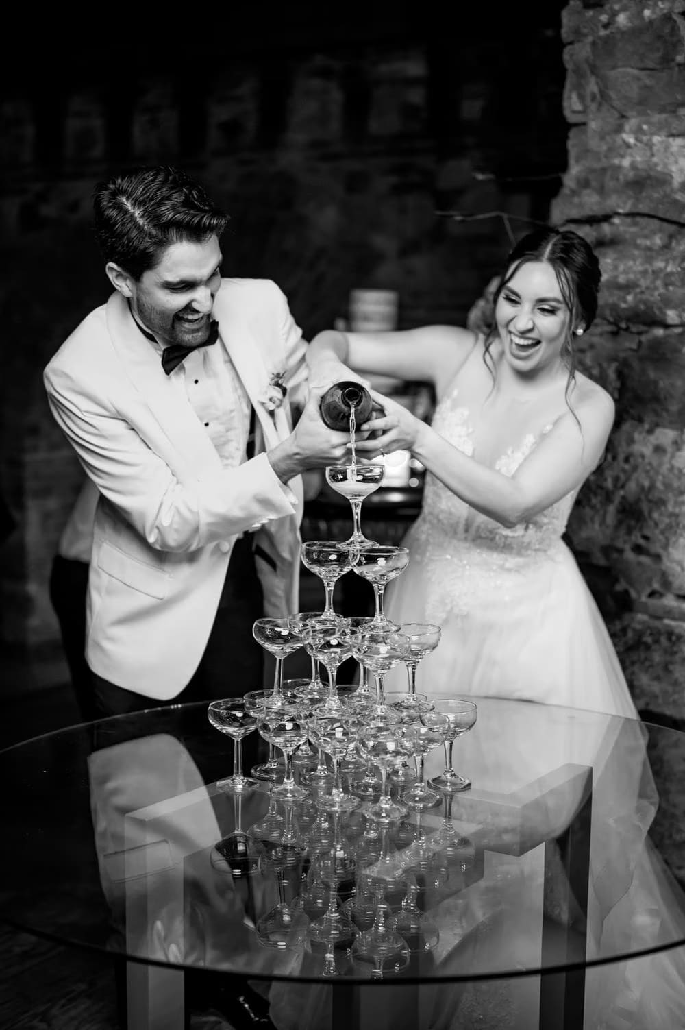 Black-and-white frame of the couple laughing as they pour the champagne tower together at the reception.