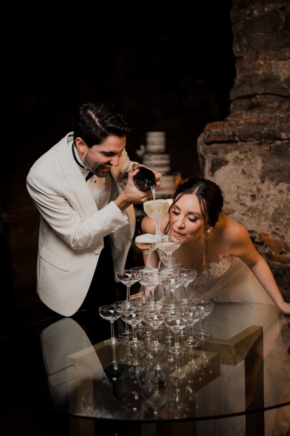 Bride sips from the top coupe of the champagne tower while the groom keeps pouring at the candle-lit bar.
